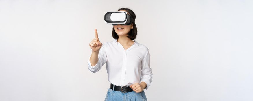 Amazed young asian woman using vr headset. Korean girl in virtual reality glasses pointing at smth and smiling, white background.