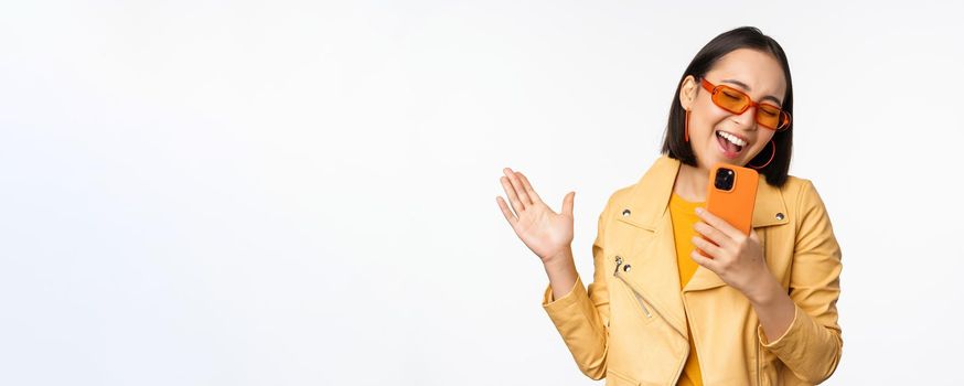 Technology and people concept. Happy dancing asian girl in sunglasses, using smartphone, singing and smiling, standing over white background.