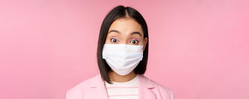 Close-up portrait of asian businesswoman in medical face mask, looking surprised, standing in suit over pink background. Copy space