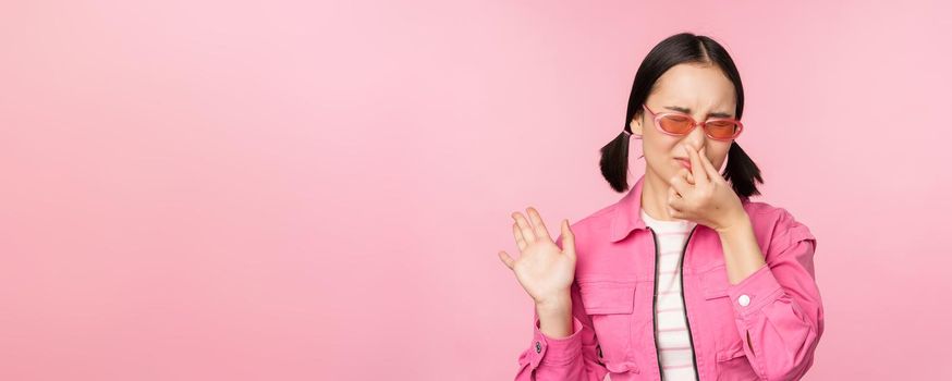 Stinky. Asian girl in sunglasses and stylish outfit, shuts her nose from disgust, dislike bad smell, standing over pink background.