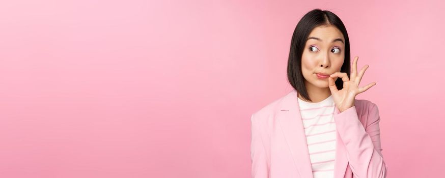 Portrait of asian corporate woman showing mouth seal, close shut lips on key gesture, promise keep secret, standing over pink background in suit.