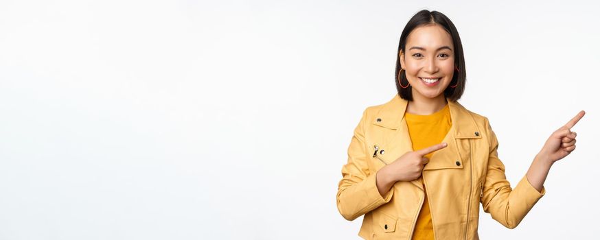 Happy asian woman smiling, pointing fingers right, inviting to check out sale, showing advertisement banner or logo, standing over white background.