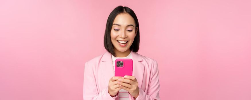 Portrait of asian girl in suit with smartphone, smiling and looking happy, standing over pink studio background. Copy space