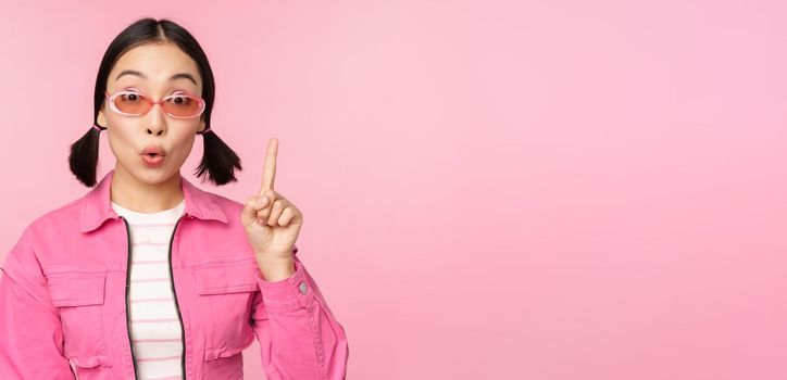 Cool and stylish asian girl, raising finger, pointing up, suggesting smth, pitching an idea or plan, standing over pink background.