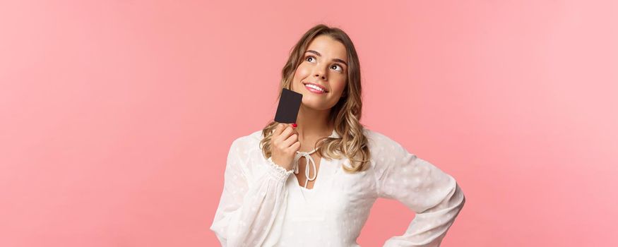 Close-up portrait of thoughtful and creative blond woman in white dress, touching chin with credit card and smiling dreamy as looking up and thinking, picturing what buy as present, pink background.