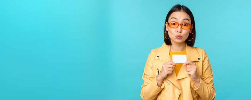 Stylish young asian woman in sunglasses, showing credit card and smiling, recommending bank, contactless payment or discounts in store, standing over blue background.