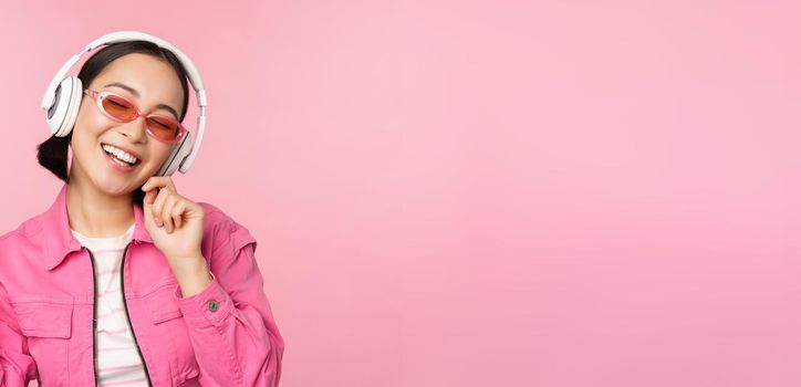 Dancing stylish asian girl listening music in headphones, posing against pink background.