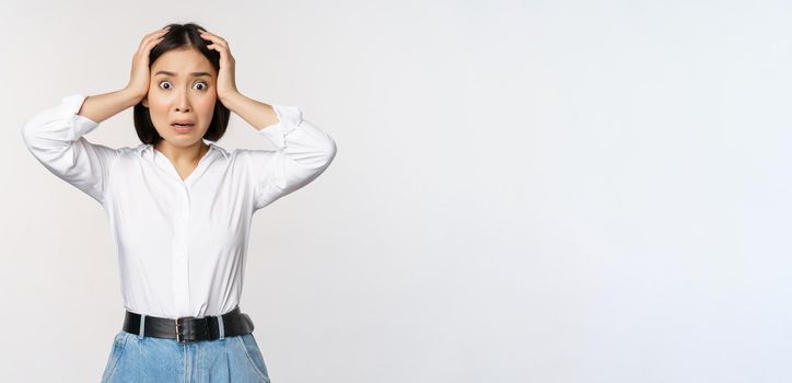 Image of shocked anxious asian woman in panic, holding hands on head and worrying, standing frustrated and scared against white background.