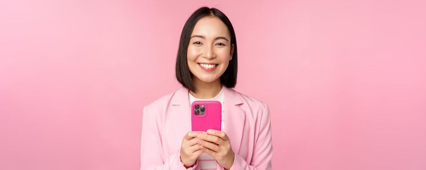 Portrait of smiling business woman, asian corporate person using smartphone, mobile phone application, standing over pink background.