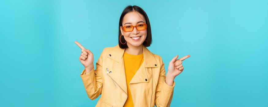 Happy stylish chinese girl in sunglasses, points fingers sideways, invites to check out, see store discount, shows left and right, stands over blue background.