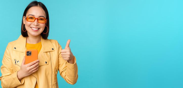 Smiling asian woman showing thumbs up, recording on mobile phone, using smartphone app and recommending it, standing over blue background.