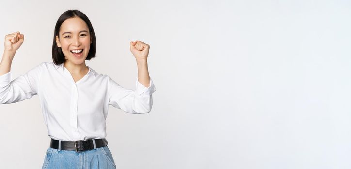 Enthusiastic asian woman rejoicing, say yes, looking happy and celebrating victory, champion dance, fist pump gesture, standing over white background.