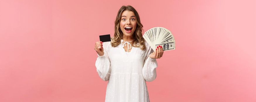 Portrait of excited happy good-looking blond girl in white dress, winning money, placed good bet, made deal, holding dollars money and credit card, smiling amused at camera, pink background.