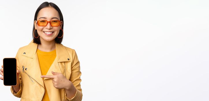 Portrait of stylish korean girl in sunglasses, smiling, pointing finger at smartphone screen, showing mobile phone application, standing over white background.