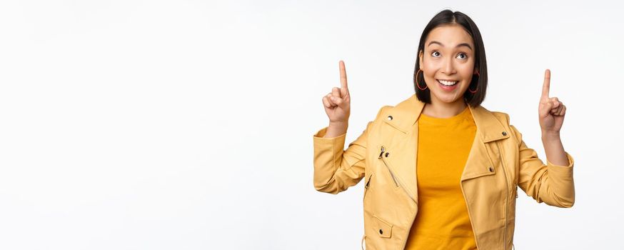 Image of smiling asian brunette woman pointing fingers up, showing advertisement with happy face, posing against white background.