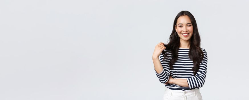 Lifestyle, people emotions and casual concept. Charming smiling korean girl in striped shirt, touching hair strand and grinning happily camera, talking to friend, white background.