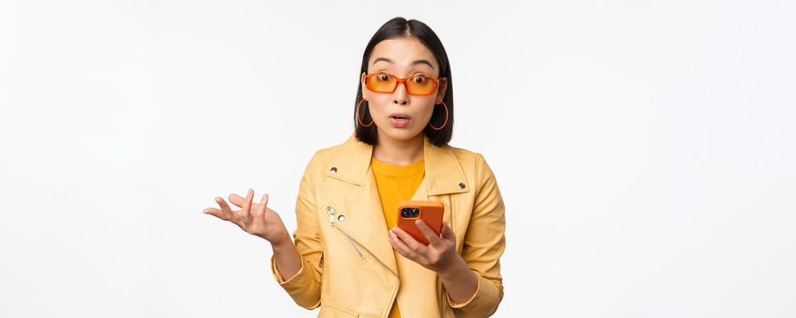 Portrait of korean girl in sunglasses, holding smartphone, looking confused and shrugging, standing over white background. Copy space