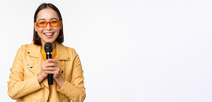 Stylish asian girl in sunglasses, singing songs with microphone, holding mic and dancing at karaoke, posing against white background.