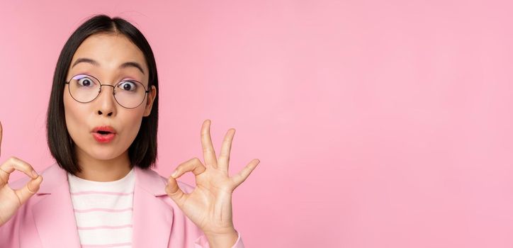 Close up portrait of business woman looks impressed and shows okay sign in approval, recommending company. Young corporate lady in glasses shows ok gesture, pink background.