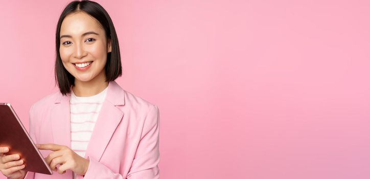 Portrait of young asian corporate woman, office lady with digital tablet, wearing suit, smiling and looking professional, posing against pink background.