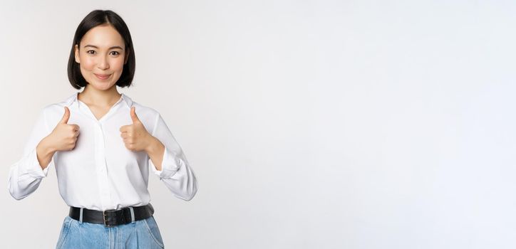 Image of beautiful adult asian woman showing thumbs up, wearing formal office, university clothing, recommending company, standing over white background.