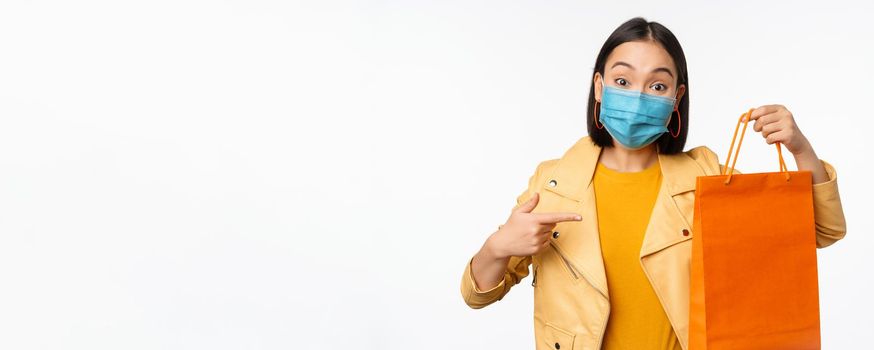 Covid-19 and shop concept. Young asian stylish woman, wearing medical face mask, holding shopping bag, going in malls during pandemic, white background.