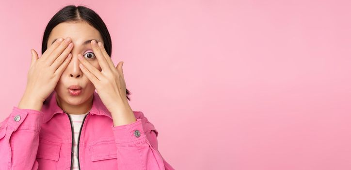 Close up portrait of young asian girl looking surprised, express amazement and wonder, peeking through fingers, standing over pink background.