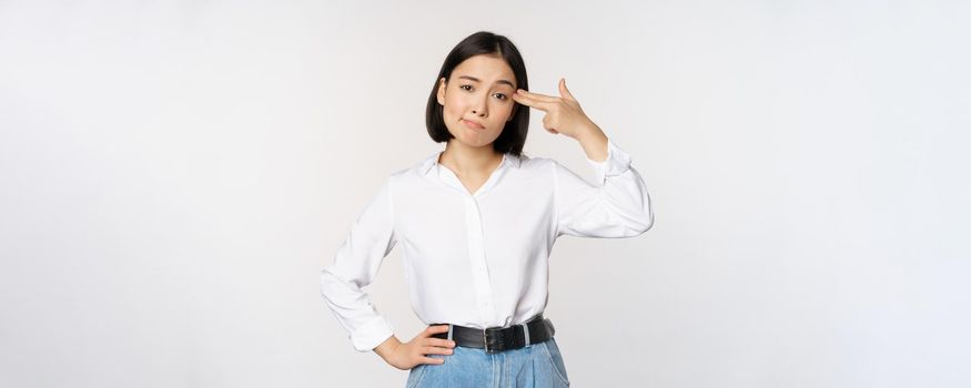 Image of tired asian woman, annoyed by smth, pointing fingers at her head, standing over white background. Copy space