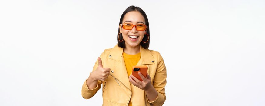 Happy stylish korean girl, wearing sunglasses, showing thumb up, holding smartphone, using mobile phone app, recommending application, online store, white background.