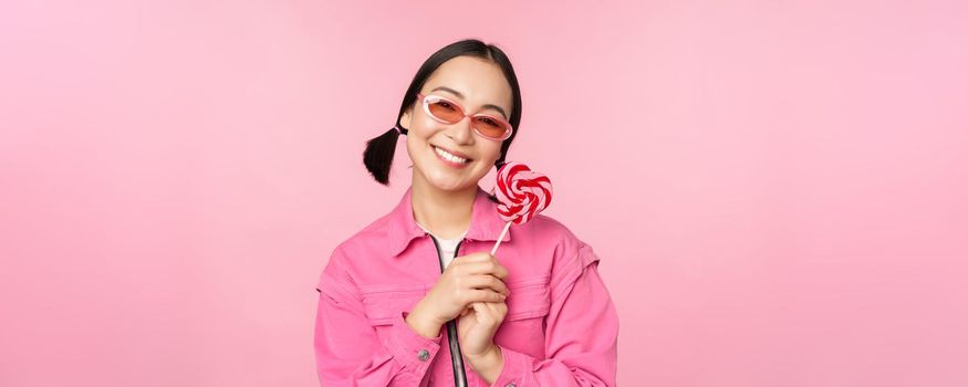 Smiling asian woman in sunglasses, holding lolipop sweets, eating candy and looking happy, standing over pink background.