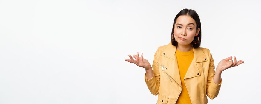 Image of confused young asian woman shrugging shoulders, looking puzzled and clueless at camera, cant understand, standing against white background.