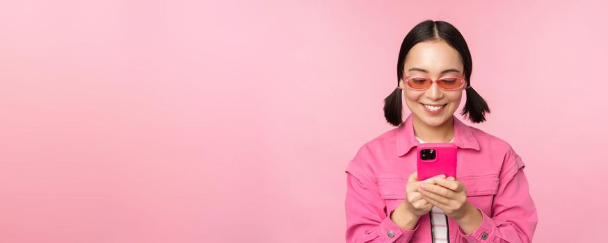 Portrait of asian girl in sunglasses using smartphone. Woman looking at mobile phone, browsing in app, standing over pink background.