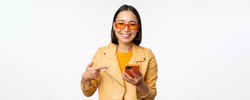 Beautiful smiling asian girl in sunglasses, pointing finger at smartphone, showing app, store on mobile phone, standing over white background.