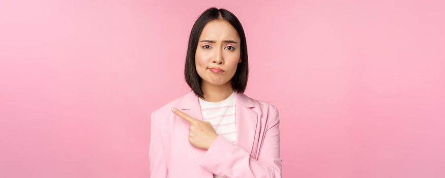 Skeptical businesswoman, pointing finger left, sulking and grimacing upset, showing smth with disappointment, standing over pink background.