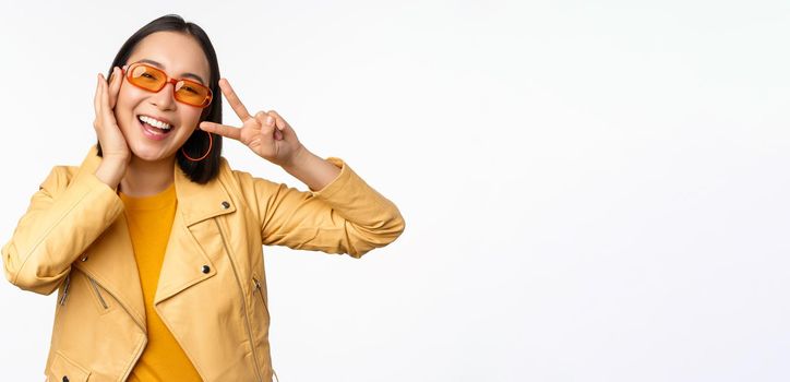 Portrait of stylish asian modern girl, wearing sunglasses and yellow jacket, showing peace, v-sign gesture, standing over white background, happy smiling face.