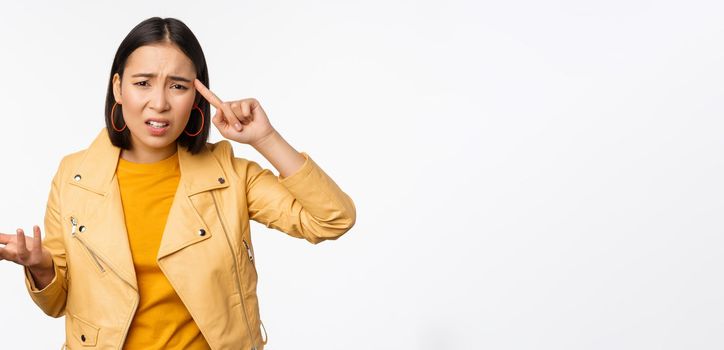 Annoyed asian woman roll finger near temple and staring frustrated, scolding someone stupid or crazy, standing over white background.