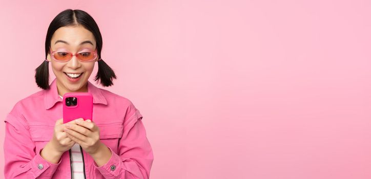 Portrait of asian girl in sunglasses using smartphone. Woman looking at mobile phone, browsing in app, standing over pink background.