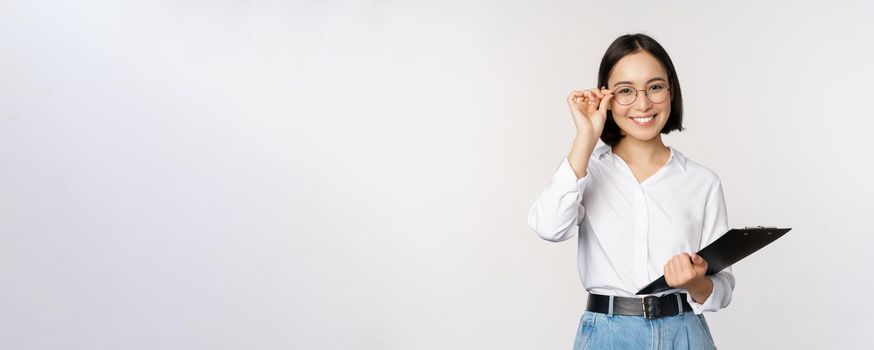 Young woman, office worker manager in glasses, holding clipboard and looking like professional, standing against white background.