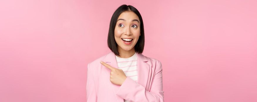 Enthusiastic young asian businesswoman, office employee pointing finger left, looking at banner, advertisement with happy smile, showing advertisement, pink background.