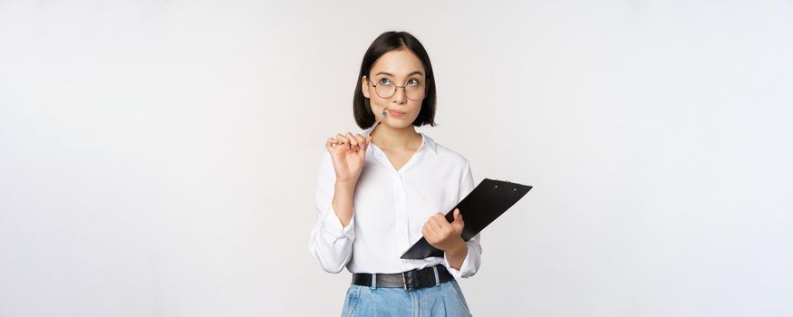 Asian girl in glasses thinks, holds pen and clipboard, writing down, making notes, standing over white background.