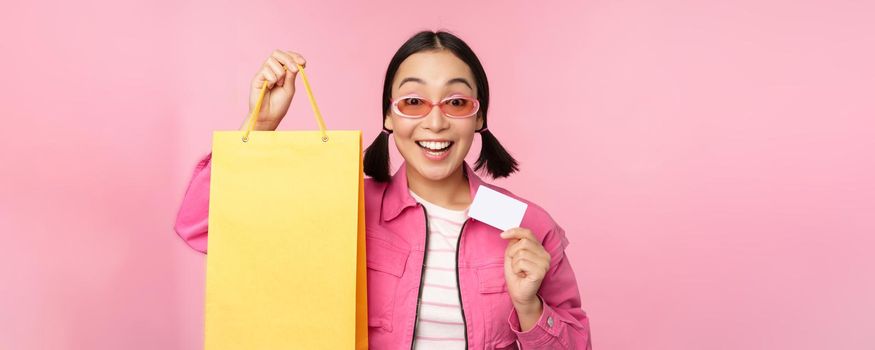 Happy young asian woman showing credit card for shopping, holding bag, buying on sale, going to the shop, store, standing over pink background.