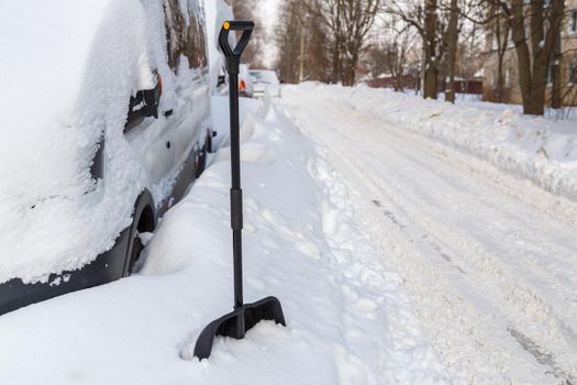 Plastic snow shovel in front of snow-covered minivan at sunny winter morning.