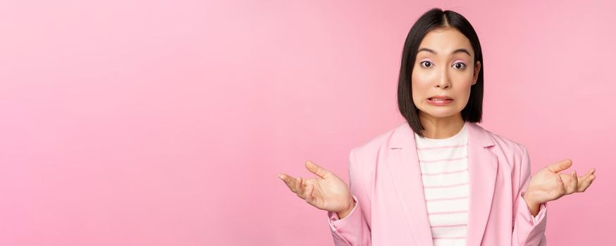 Portrait of confused asian businesswoman shrugging shoulders, looking clueless and puzzled, dont know, cant say, standing over pink background in office suit.