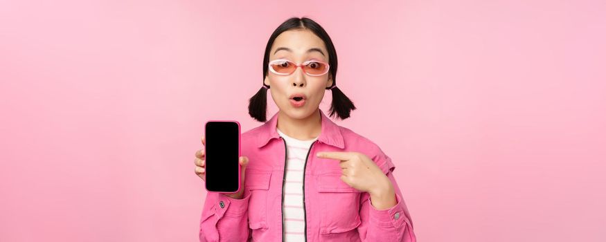Enthusiastic asian woman in stylish clothes, sunglasses, pointing finger at mobile phone screen, showing smartphone application, standing over pink background.