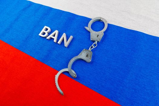 the word ban laid with silver metal letters near opened handcuffs over fabric tricolor flag of the Russian Federation