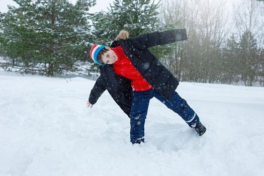 A cheerful teenager in a dark jacket and a red sweater stands in a winter park on the snow, spreading his arms and balancing