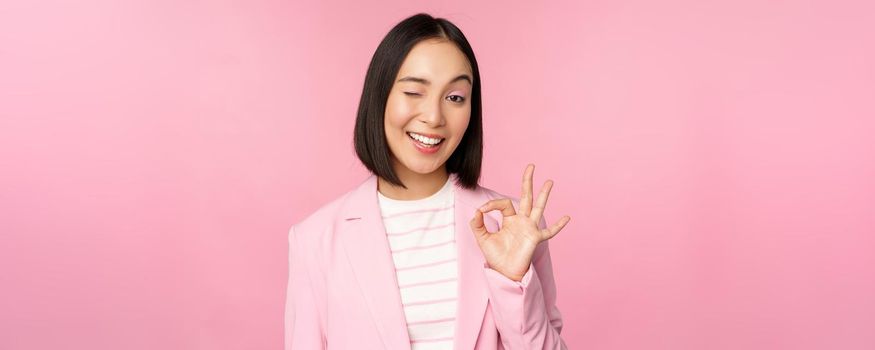 Excellent work, well done gesture. Smiling asian businesswoman showing okay, ok sign, praise good work, recommending company, looking as confident professional, pink background.