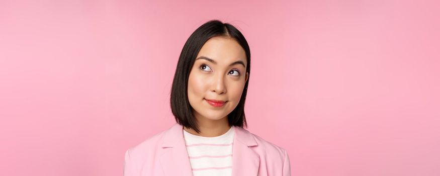 Close up portrait of young asian businesswoman thinking, smiling thoughtful and looking at upper left corner, standing over pink background.