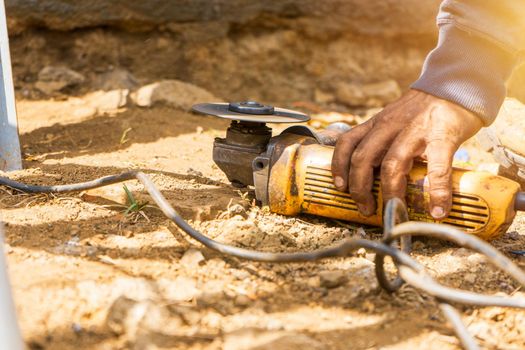 Closeup of a hand of a blue collar worker from Latin America grabbing a compact electric handheld circular saw that is on the dirt floor.