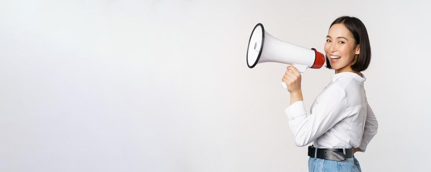 Beautiful young asian woman talking in megaphone, screams in speakerphone and smiling, making announcement, shout out information, standing over white background.